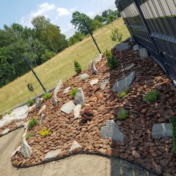 Hubert Kamiński - Aranżacja ogrodu z korą, kamieniami i niskimi roślinami iglastymi, widok z boku, ogrodzenie panelowe w tle, słoneczny dzień.