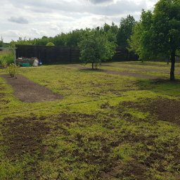Hubert Kamiński - Przygotowany teren pod trawnik: wyrównana ziemia z widocznymi śladami po pracach ziemnych, drzewa i krzewy w tle, ogrodzenie z siatki i drewniany płot.