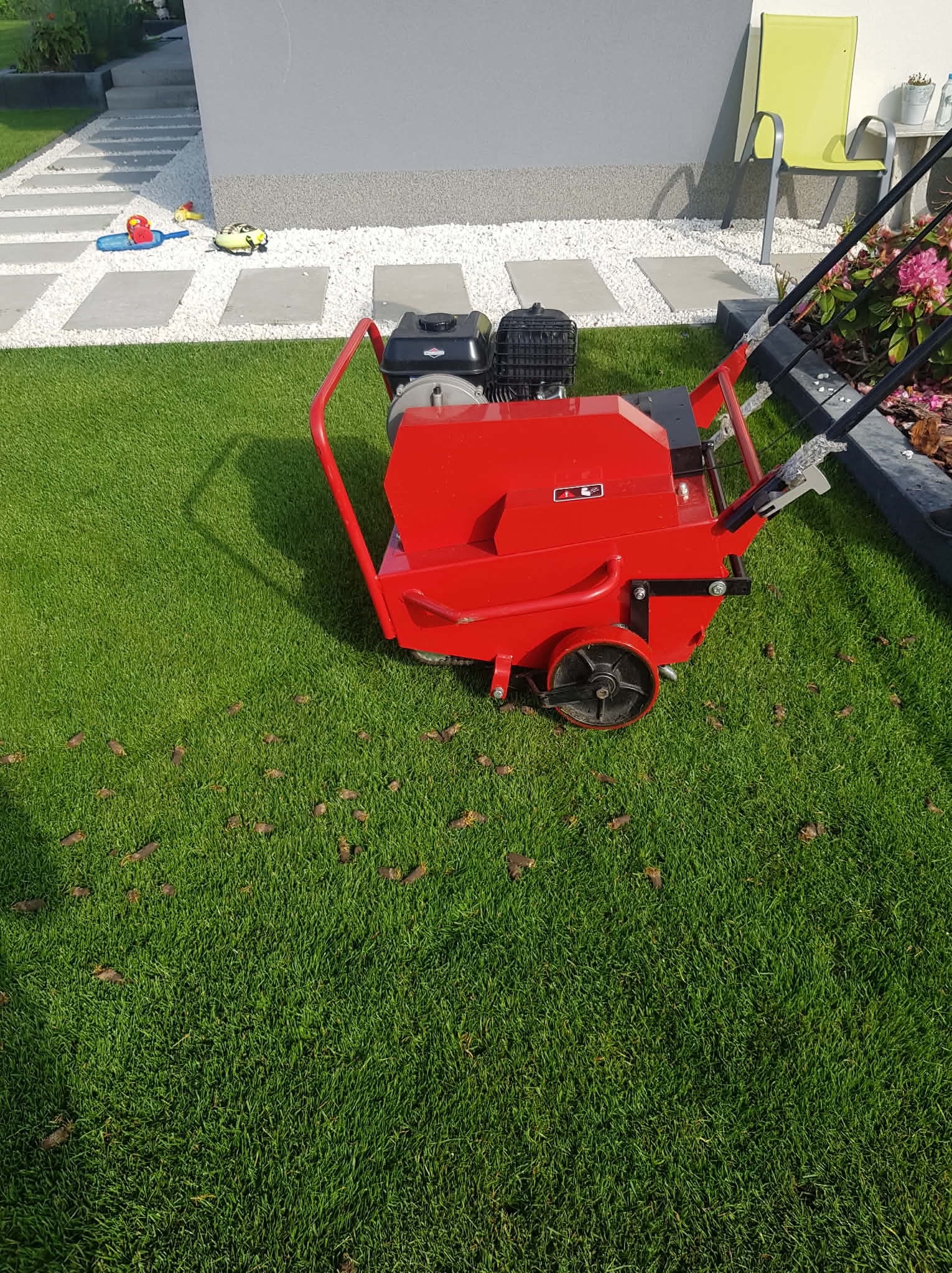 Czerwony aerator do trawy na zadbanym trawniku z widocznymi śladami po aeracji, obok ścieżka z białego kamienia i zabawki dziecięce. Trawa gęsta, zielona, po zabiegu pielęgnacyjnym.