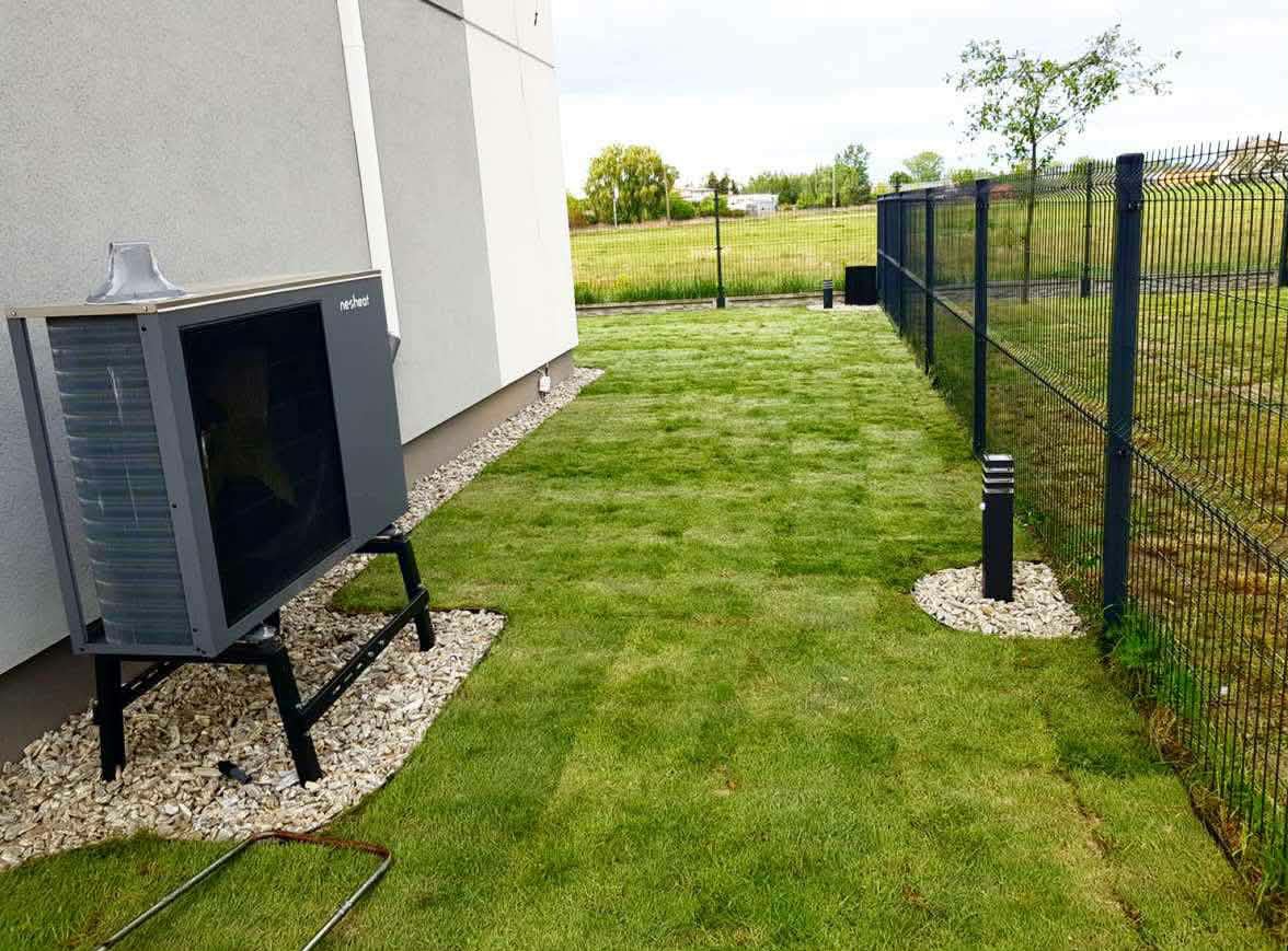 Świeżo założony trawnik z rolki przy nowoczesnym domu, z widoczną pompą ciepła i oświetleniem ogrodowym wzdłuż ogrodzenia. Ujęcie z poziomu gruntu, podkreślające gęstość trawy.