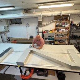 KAPITAN REKLAMA SPÓŁKA Z OGRANICZONĄ ODPOWIEDZIALNOŚCIĄ - Pracownik warsztatu łączy elementy przestrzennej litery reklamowej na stole roboczym, w tle narzędzia i półki z akcesoriami.