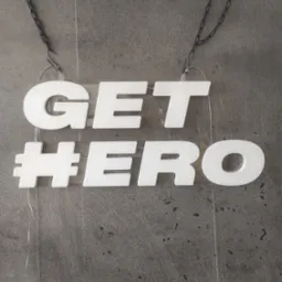 Biały neonowy napis 'GET #HERO' zawieszony na czarnym łańcuszku, widoczny na tle szarej ściany. Nowoczesny design, wyraźny kontrast, atrakcyjny element dekoracyjny.