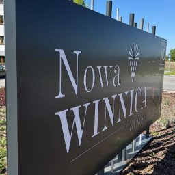 KAPITAN REKLAMA SPÓŁKA Z OGRANICZONĄ ODPOWIEDZIALNOŚCIĄ - Czarny szyld z białym napisem 'Nowa Winnica Miekinta' i symbolem winogron, umieszczony na zewnątrz budynku w słoneczny dzień. Szyld wsparty na metalowych słupkach.
