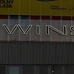 KAPITAN REKLAMA SPÓŁKA Z OGRANICZONĄ ODPOWIEDZIALNOŚCIĄ - Podświetlany neonowy napis 'TWINS' na budynku z banerem 'Dolny Śląsk'. Neon ma kremowe światło, widoczne elementy konstrukcyjne i tło budynku.