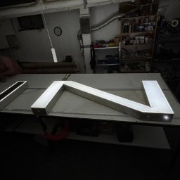 KAPITAN REKLAMA SPÓŁKA Z OGRANICZONĄ ODPOWIEDZIALNOŚCIĄ - Metalowa, podświetlana konstrukcja w kształcie liter 'IN' na stole w warsztacie we Wrocławiu. W tle półki z narzędziami i osoba pracująca przy stole.