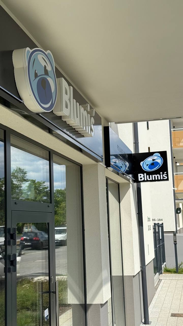 Zewnętrzny szyld reklamowy z logo i nazwą firmy 'Blumiś' nad wejściem do lokalu. Szyld z wizerunkiem misia i napisem w białych, przestrzennych literach.