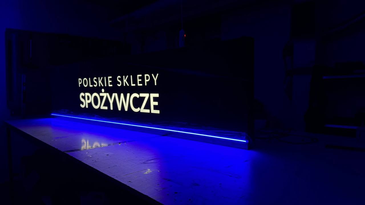 Podświetlany szyld z napisem 'Polskie Sklepy Spożywcze', oświetlony niebieskim światłem LED, widoczny z perspektywy lekko z dołu, na ciemnym tle.