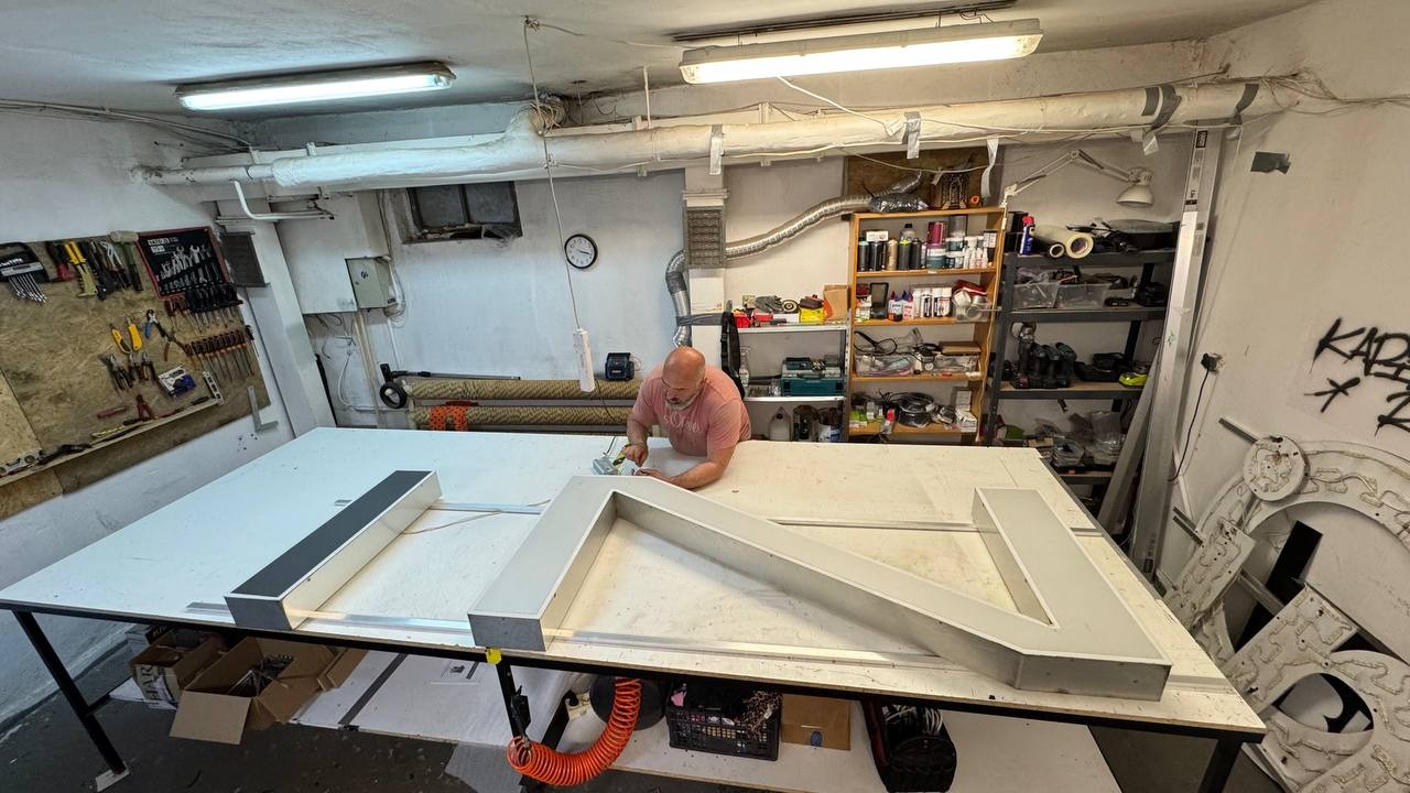 Pracownik warsztatu łączy elementy przestrzennej litery reklamowej na stole roboczym, w tle narzędzia i półki z akcesoriami.