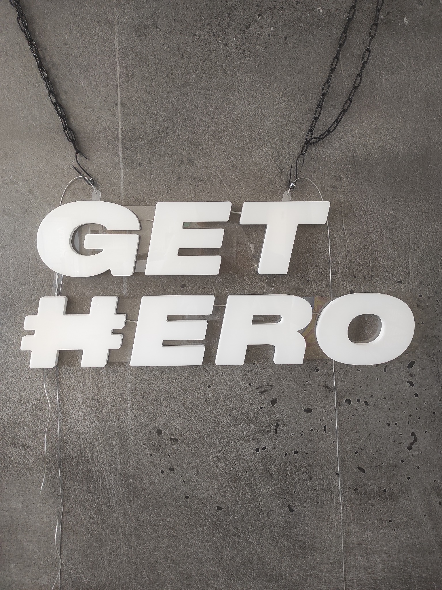 Biały neonowy napis 'GET #HERO' zawieszony na czarnym łańcuszku, widoczny na tle szarej ściany. Nowoczesny design, wyraźny kontrast, atrakcyjny element dekoracyjny.