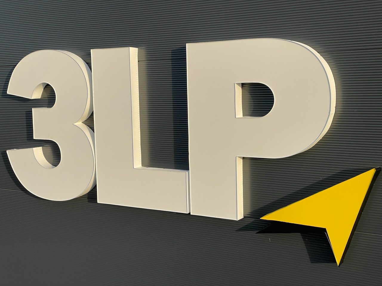 Zewnętrzny szyld 3LP z żółtą strzałką na szarej, poziomo żebrowanej elewacji, widoczny w ukośnym ujęciu z dołu, w jasnym oświetleniu.