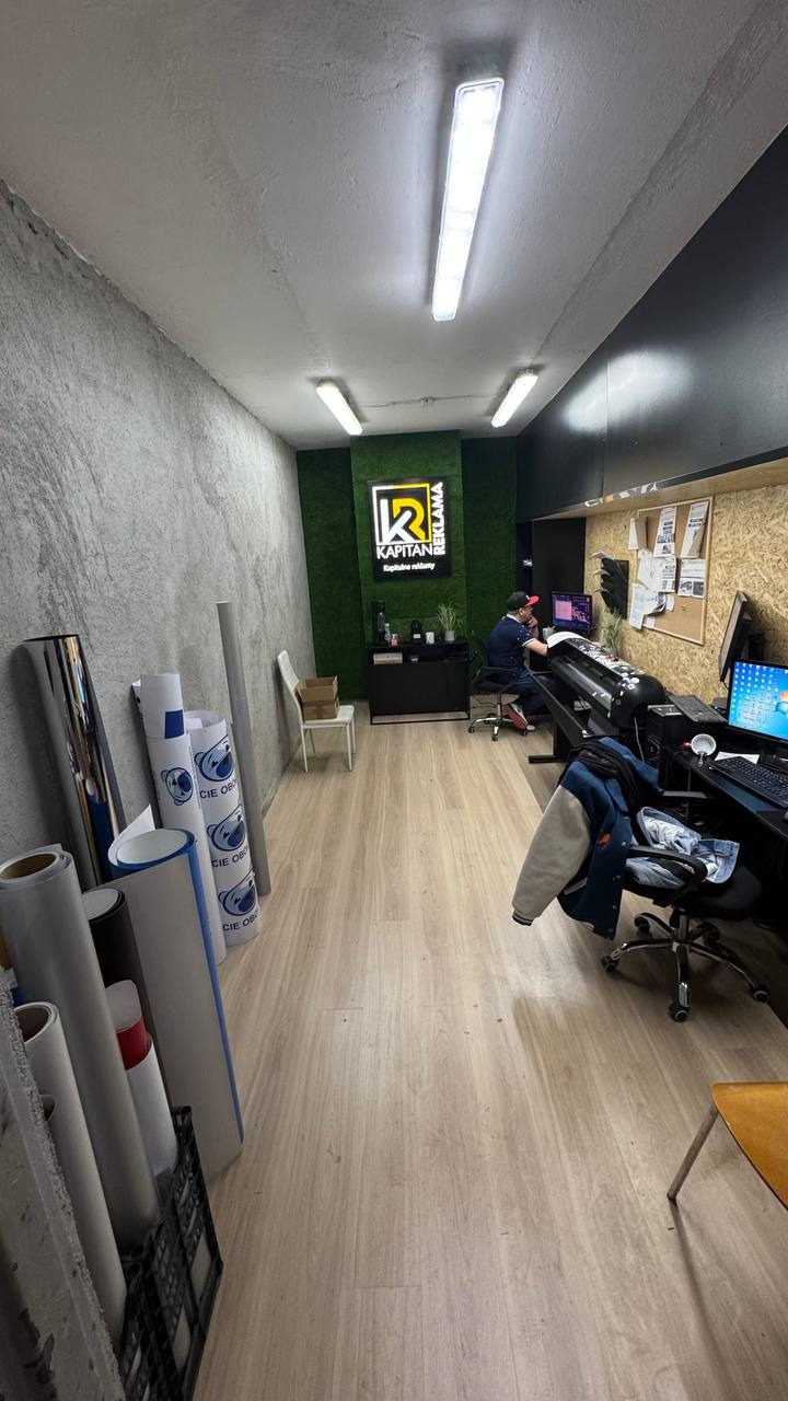 Wnętrze biura firmy reklamowej: rolki folii, ploter, biurko z komputerem, pracownik przy pracy i podświetlone logo firmy na ścianie z mchu.