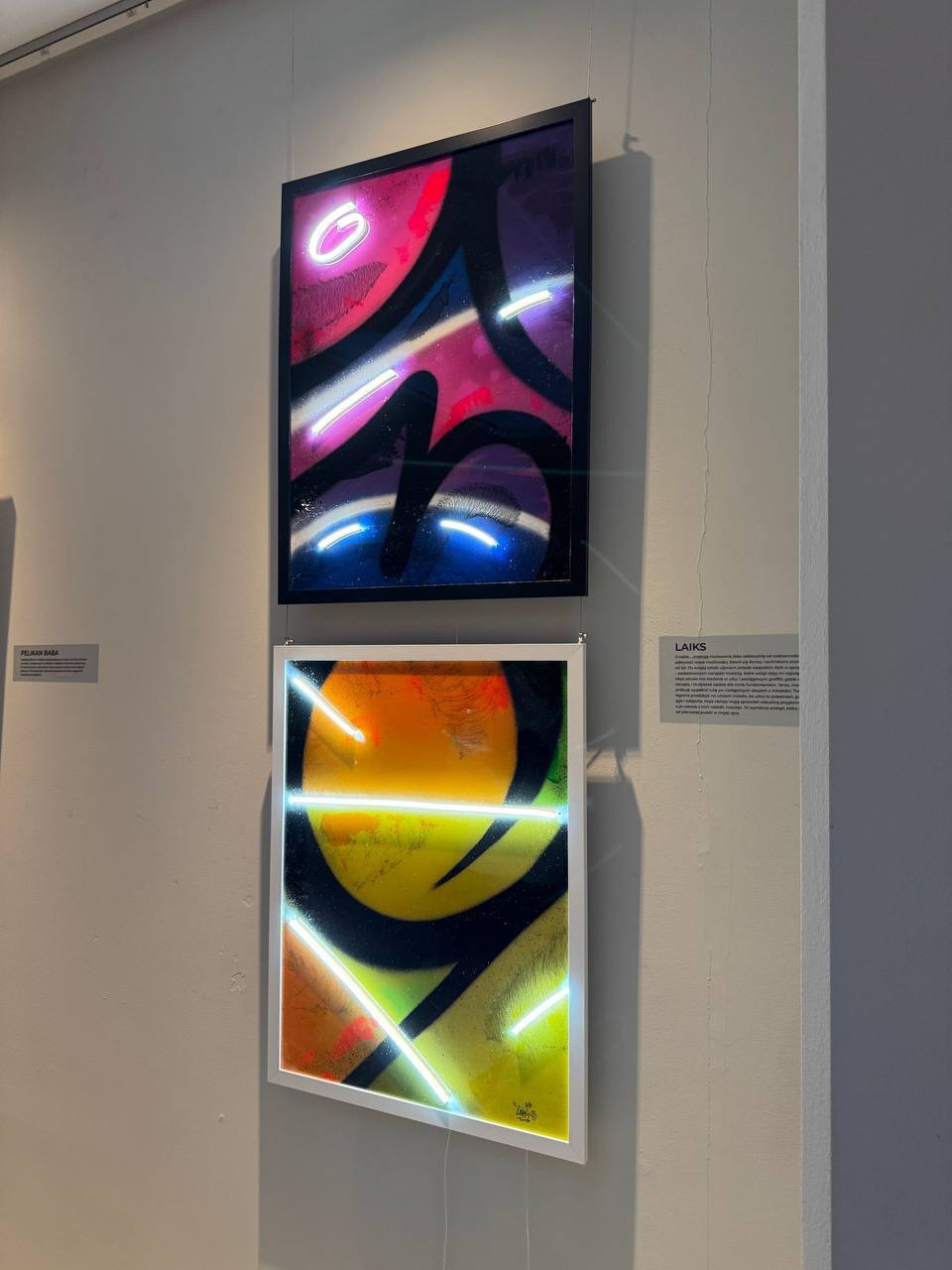 Dwa oprawione obrazy graffiti na ścianie galerii: górny w odcieniach różu i fioletu, dolny w żółci i pomarańczy, oba z neonowymi akcentami.