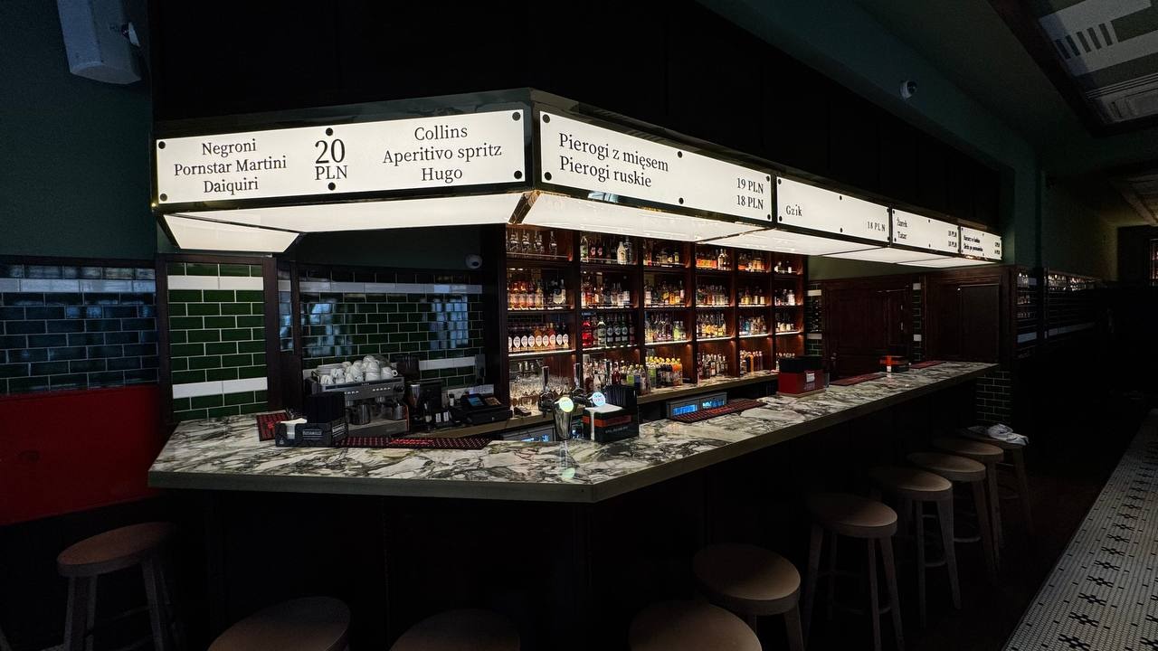 Wnętrze baru z marmurowym blatem, rzędem hokerów i podświetlonym menu z drinkami i pierogami. Na półkach za barem widoczne butelki alkoholi i ekspres do kawy.