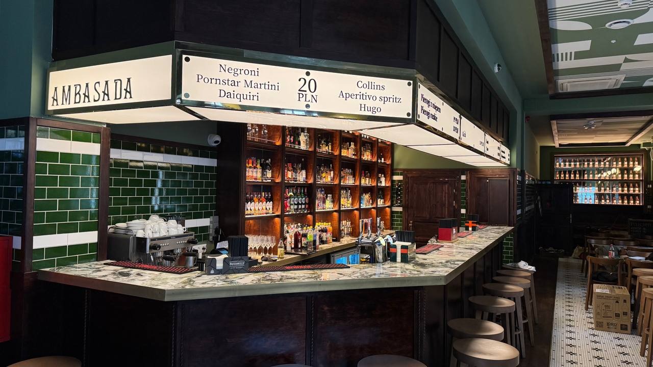 Wnętrze baru 'Ambasada' z zielonymi płytkami, marmurowym blatem i podświetlanymi półkami z alkoholem. Nad barem menu drinków z cenami.