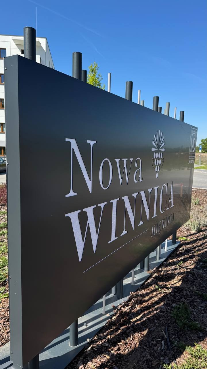 Czarny szyld z białym napisem 'Nowa Winnica Miekinta' i symbolem winogron, umieszczony na zewnątrz budynku w słoneczny dzień. Szyld wsparty na metalowych słupkach.