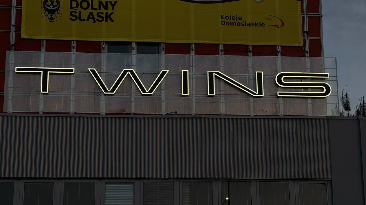 Podświetlany neonowy napis 'TWINS' na budynku z banerem 'Dolny Śląsk'. Neon ma kremowe światło, widoczne elementy konstrukcyjne i tło budynku.