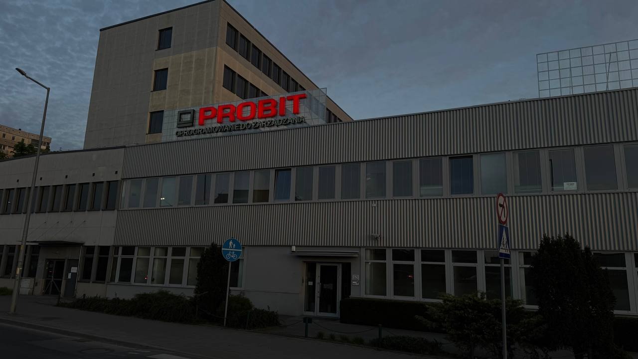 Budynek biurowy firmy Probit z czerwonym logotypem na fasadzie, widoczny z poziomu ulicy w pochmurny dzień. Charakterystyczna architektura lat 90.