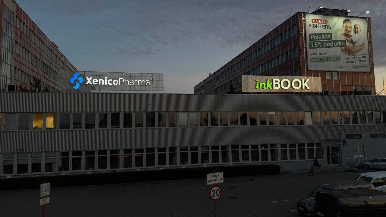 Budynek biurowy z logo firm XenicoPharma i inkBOOK na dachu. Billboard Cancer Fighters z prośbą o przekazanie 1,5% podatku. Widok z parkingu.