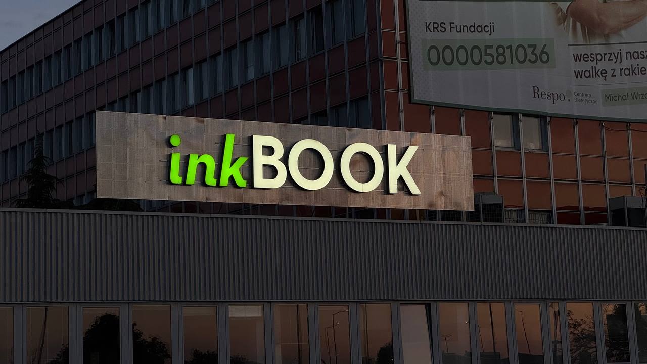 Zewnętrzny szyld firmy InkBOOK z zielonym napisem 'ink' i białym 'BOOK' na tle budynku biurowego z billboardem fundacji KRS.