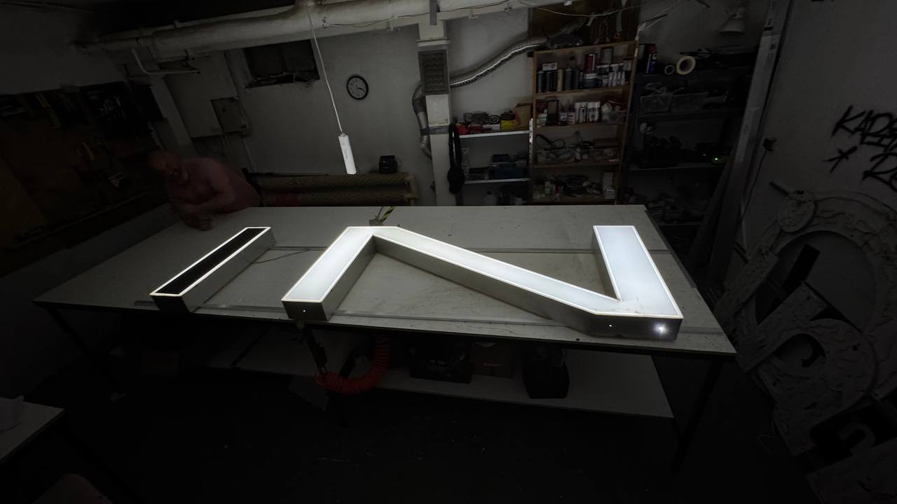 Metalowa, podświetlana konstrukcja w kształcie liter 'IN' na stole w warsztacie we Wrocławiu. W tle półki z narzędziami i osoba pracująca przy stole.