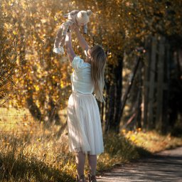 Adrian Ekiert Fotografia - Mama w jasnej spódnicy podnosi w górę uśmiechnięte dziecko w pluszowym kombinezonie na tle drzew z żółtymi liśćmi w słoneczny dzień.