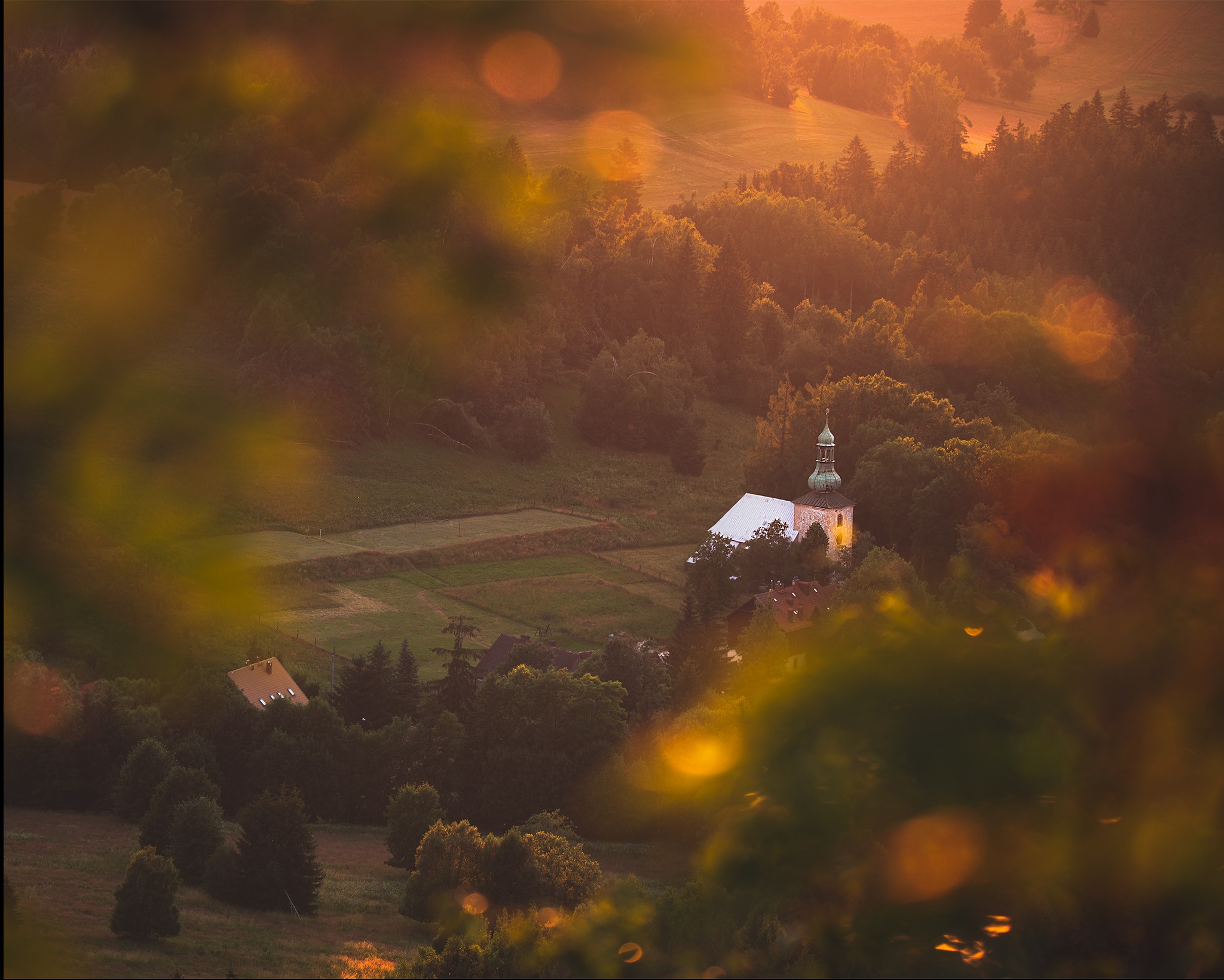 Pejzaż z kościołem w oddali, ujęty przez liście z efektem bokeh, ciepłe światło zachodzącego słońca, malowniczy krajobraz, wiejska sceneria.