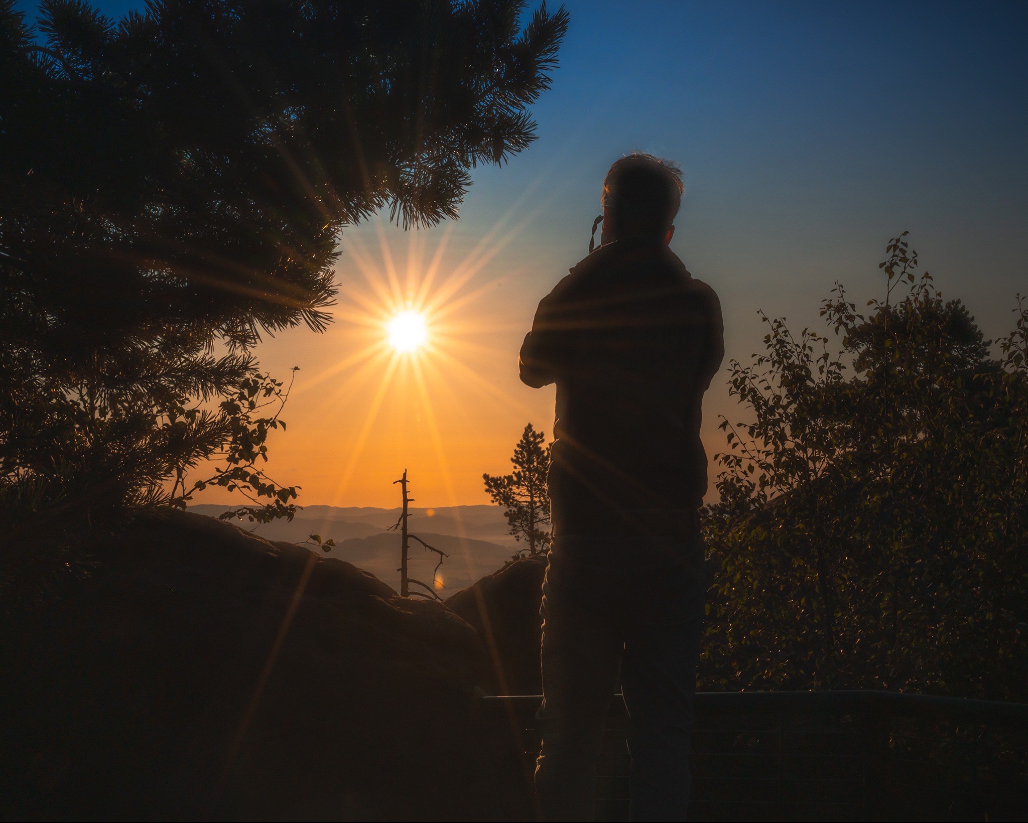 Sylwetka osoby podziwiającej wschód słońca z górskim krajobrazem w tle, uchwycona z niskiego kąta, z elementami roślinności w kadrze.