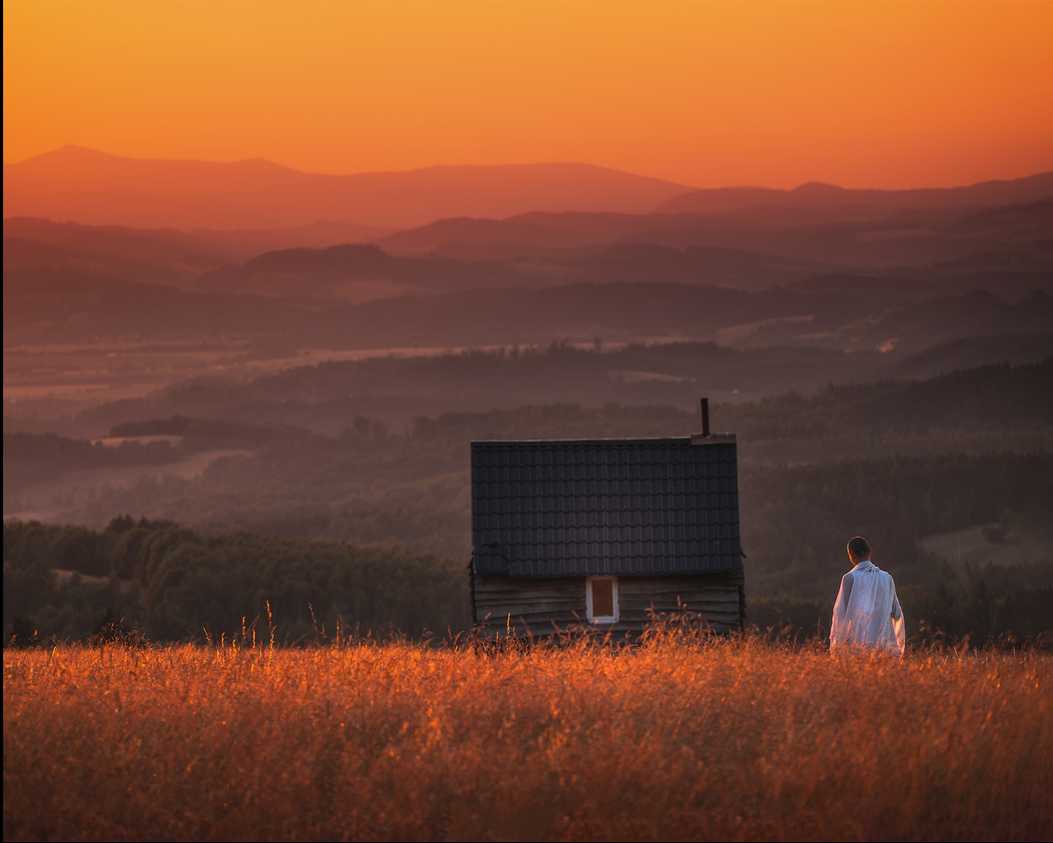 Pejzaż z Kłodzka o zachodzie słońca: sylwetka osoby w polu, mały budynek i góry w tle, ciepłe barwy, spokojna atmosfera. Profesjonalna fotografia.