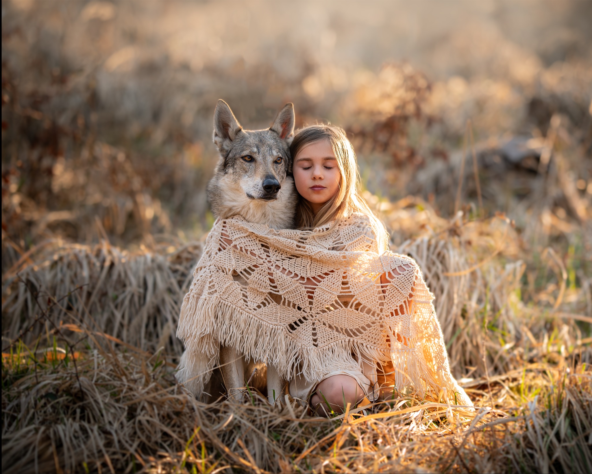 Dziewczynka z zamkniętymi oczami, przytulona do wilka, okryta beżowym, ażurowym kocem z frędzlami, na tle łąki w ciepłym świetle.