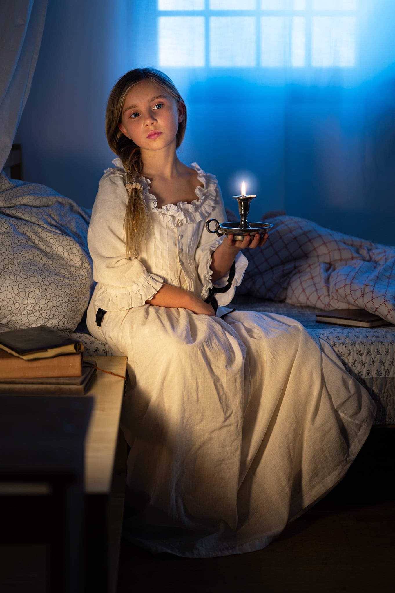 Dziewczynka w białej sukience siedzi na łóżku, trzymając świecę w świeczniku. Za nią widoczne okno z niebieskim światłem. Klimatyczne oświetlenie świecą.