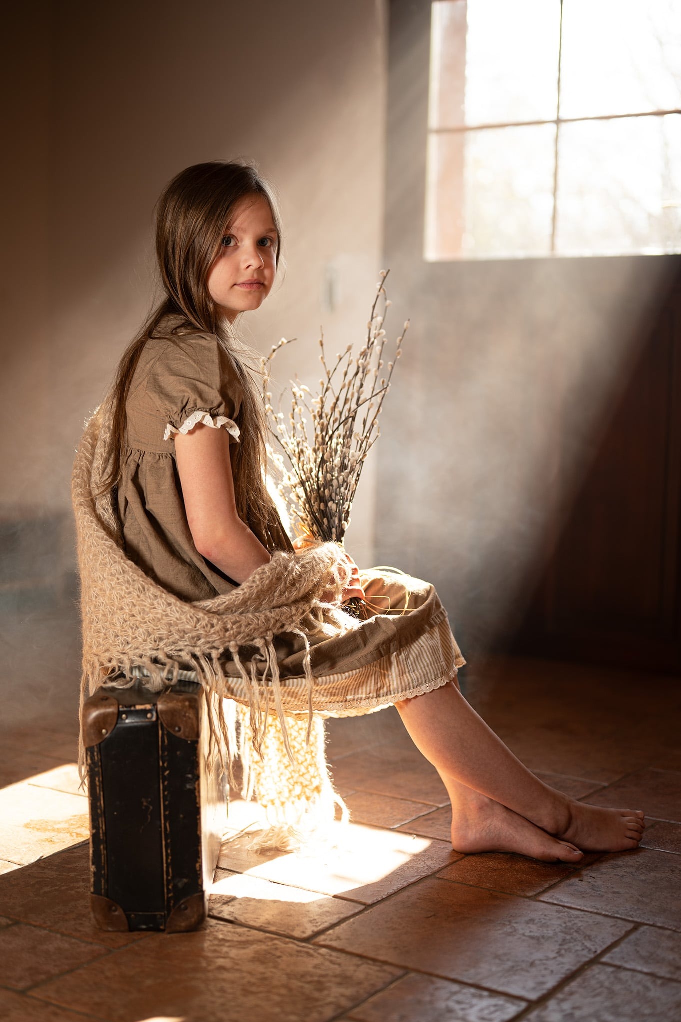 Dziewczynka w brązowej sukience siedzi na starej walizce, trzymając bukiet bazi. Ciepłe światło słoneczne pada z okna, tworząc nastrojową scenerię we wnętrzu.