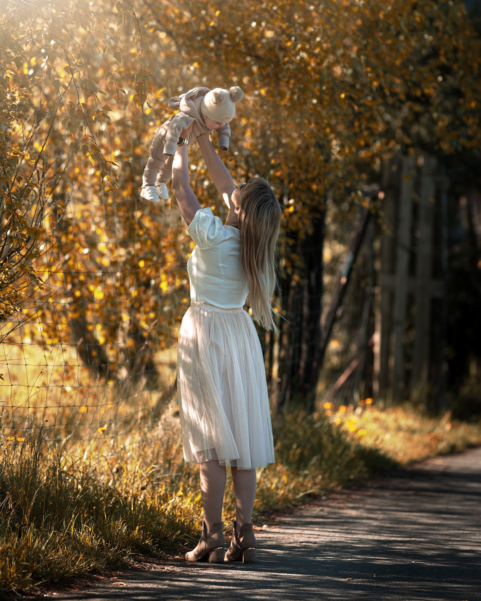 Mama w jasnej spódnicy podnosi w górę uśmiechnięte dziecko w pluszowym kombinezonie na tle drzew z żółtymi liśćmi w słoneczny dzień.