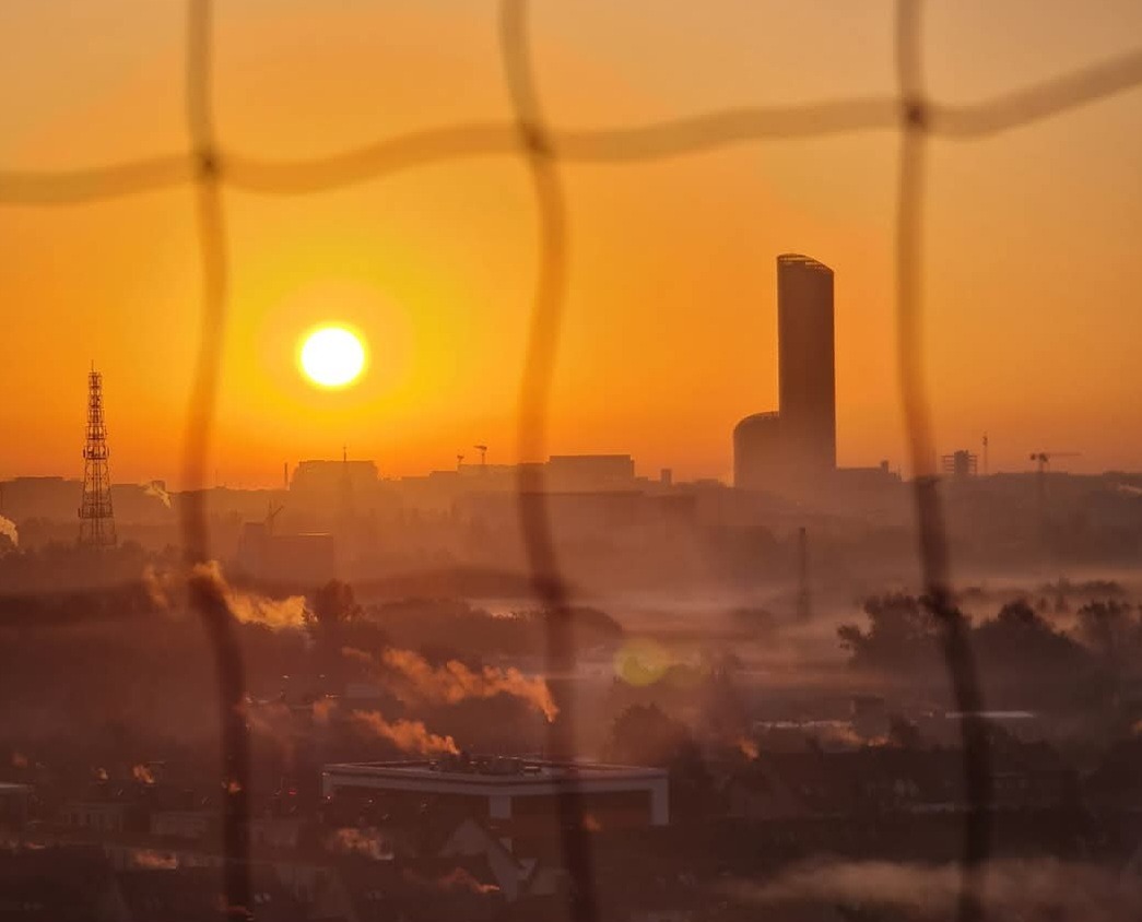 Pomarańczowy wschód słońca nad miastem, widoczny przez siatkę. Budynki i wieże w tle spowite mgłą, nadając scenerii eteryczny charakter.