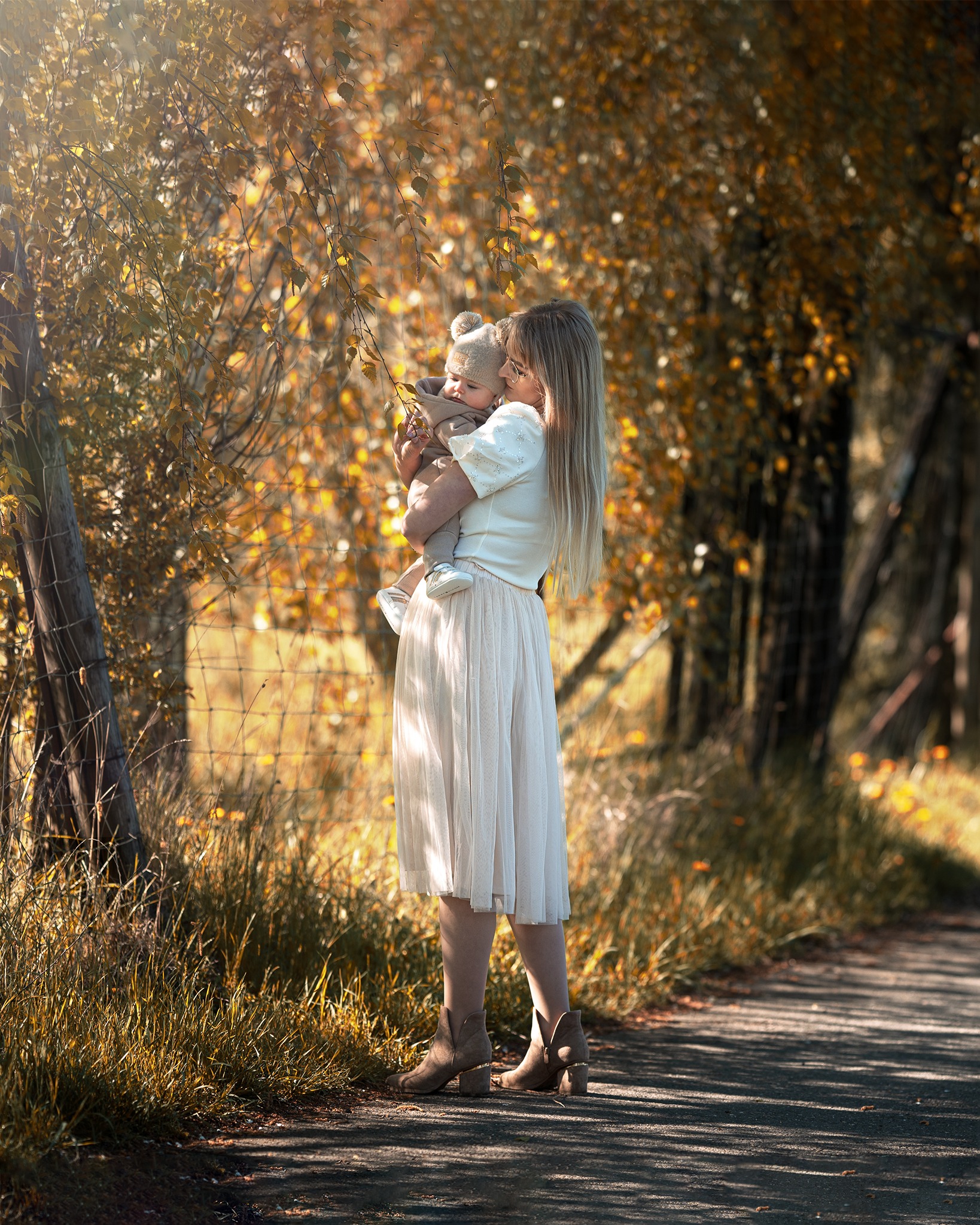 Jesienna sesja rodzinna: Matka w zwiewnej spódnicy trzyma na rękach niemowlę w czapce z pomponem, oboje pod drzewem z żółtymi liśćmi, ciepłe światło.