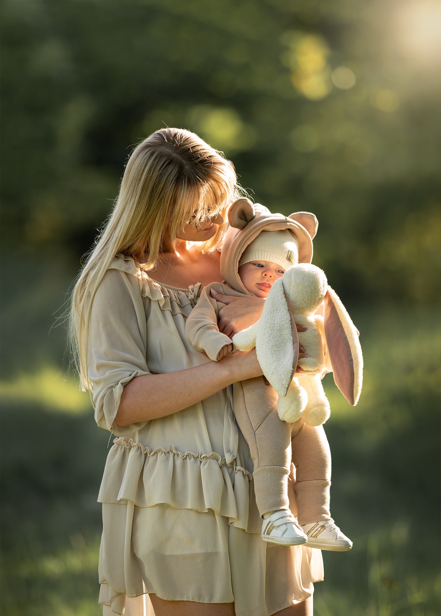 Mama z blond włosami trzyma dziecko w beżowym kombinezonie z uszami i pluszowym królikiem na tle zieleni, w miękkim, naturalnym świetle.