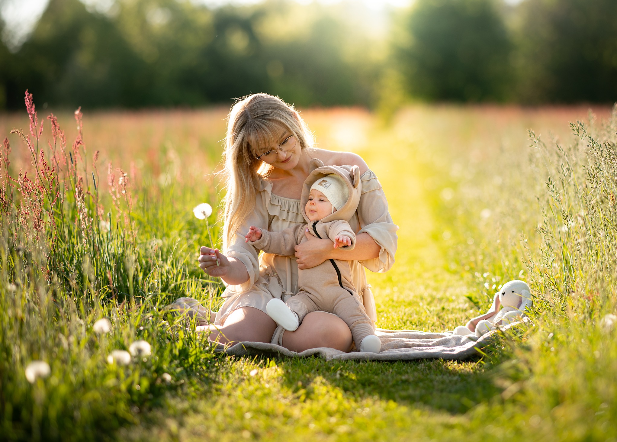 Mama w okularach i dziecko w beżowym kombinezonie siedzą na kocu w trawie, trzymając dmuchawca. Z boku leży pluszowy królik, a w tle rozmyty krajobraz.