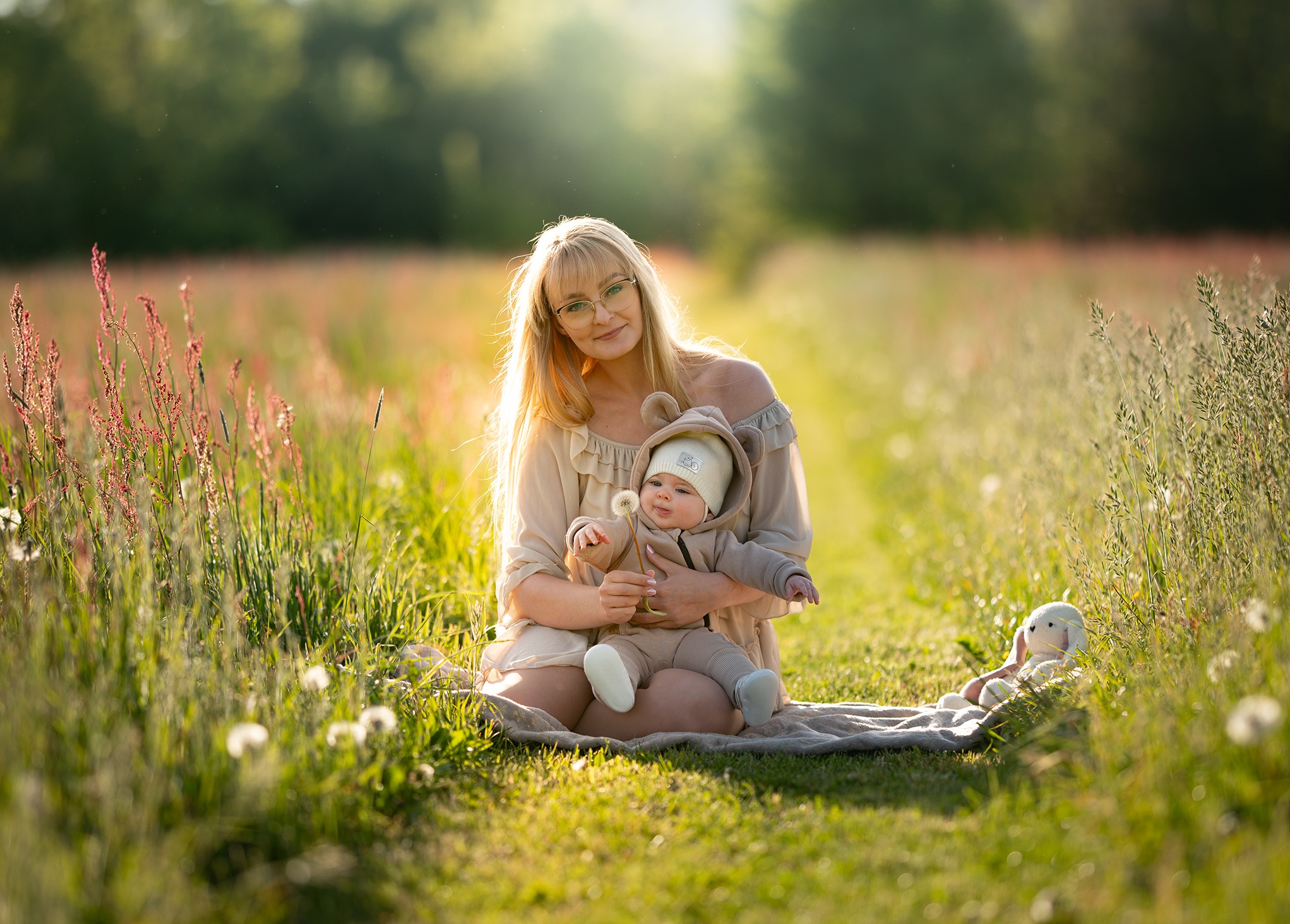 Mama z uśmiechniętym niemowlęciem w beżowym ubranku siedzą na kocu na łące, trzymając dmuchawca. W tle rozmyte trawy i drzewa, obok pluszowy królik.