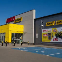 Fasada sklepu Brico Marche z żółtą strefą wejściową i szarymi ścianami. Widoczne reklamy produktów ogrodniczych i budowlanych oraz miejsca parkingowe dla niepełnosprawnych.