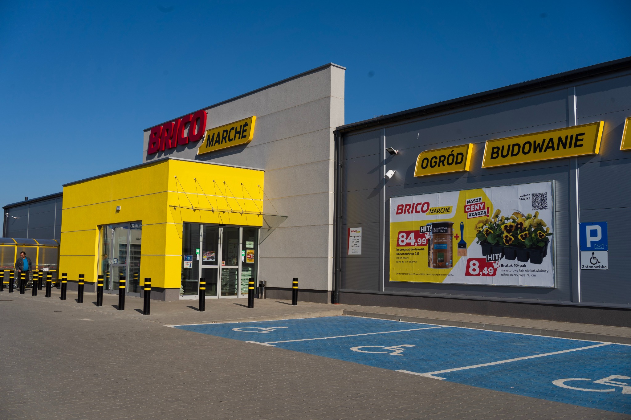 Fasada sklepu Brico Marche z żółtą strefą wejściową i szarymi ścianami. Widoczne reklamy produktów ogrodniczych i budowlanych oraz miejsca parkingowe dla niepełnosprawnych.