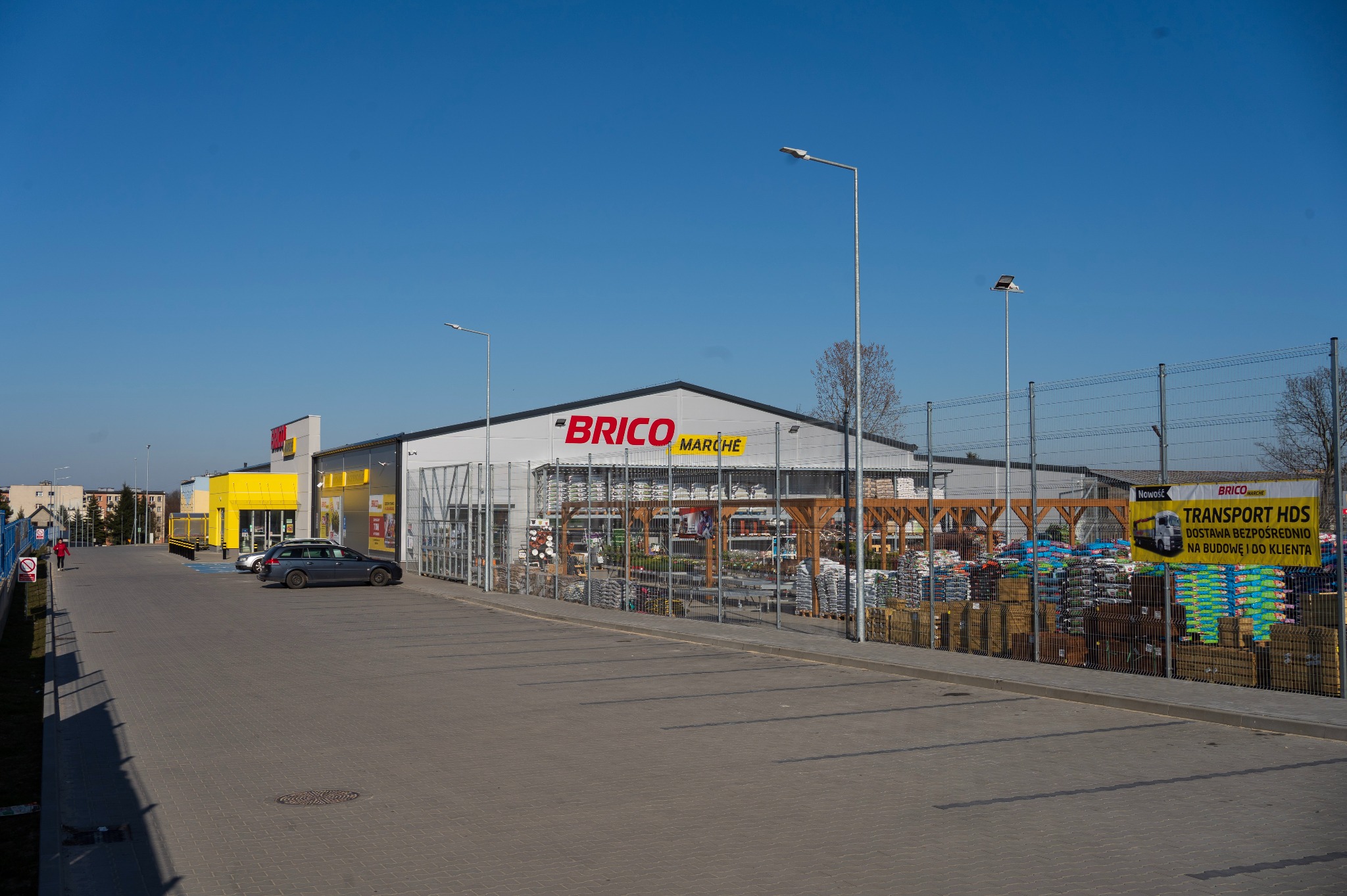Szeroki kadr na sklep budowlany Brico Marche z parkingiem, prezentujący ofertę materiałów i konstrukcji ogrodowych na zewnątrz, pod błękitnym niebem.