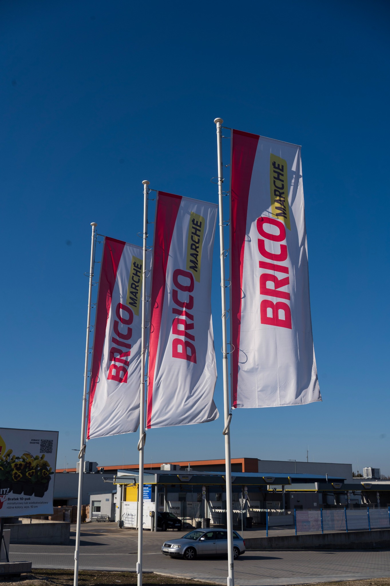 Trzy białe flagi z logo Brico Marche na tle błękitnego nieba, widoczne budynki sklepu i zaparkowany samochód, reklama bratków po lewej stronie.