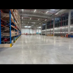 Rozległa hala magazynowa z regałami wypełnionymi towarami, posadzka betonowa. W oddali widoczni pracownicy w odblaskowych ubraniach.