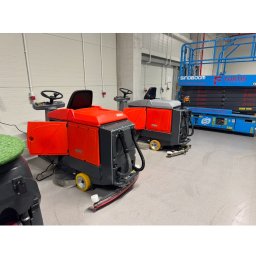 Agma Trade sp zoo - Dwie pomarańczowe maszyny czyszczące Hako w hali przemysłowej. W tle podnośnik Sinoboom Fortis. Widoczne ślady czyszczenia podłogi. Hala oświetlona lampami.