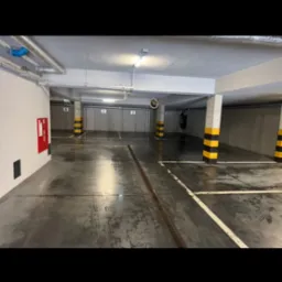 Wnętrze parkingu podziemnego z betonową posadzką, słupami w pasy żółto-czarne i numerowanymi miejscami. Na posadzce widoczne ślady wilgoci.