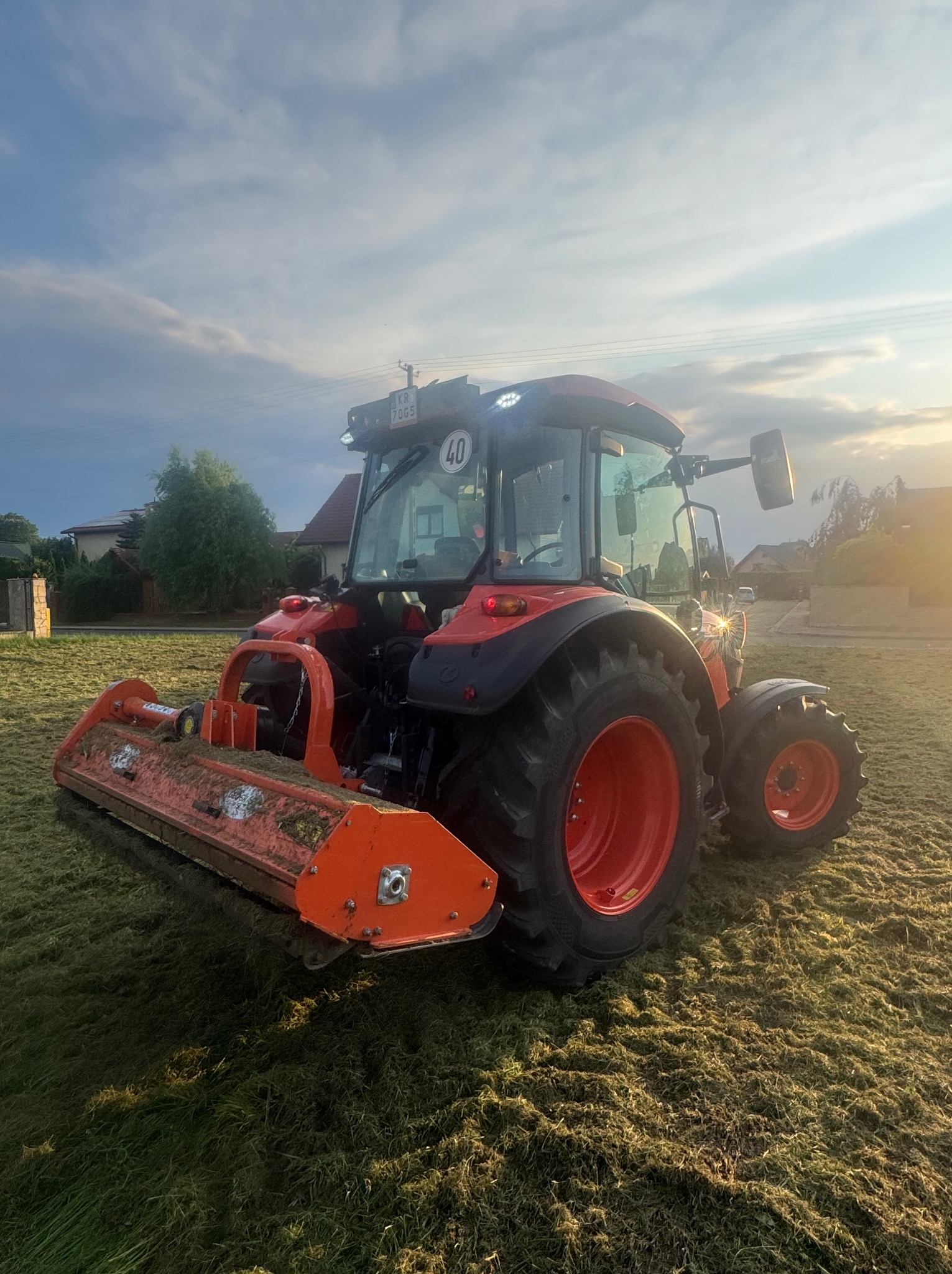 Czerwony traktor po koszeniu trawy na polu, widoczne ślady cięcia, w tle domy i drzewa, słoneczny dzień.