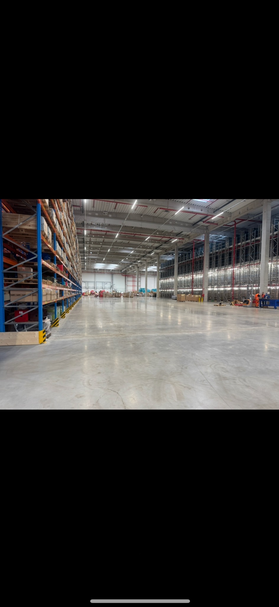 Rozległa hala magazynowa z regałami wypełnionymi towarami, posadzka betonowa. W oddali widoczni pracownicy w odblaskowych ubraniach.