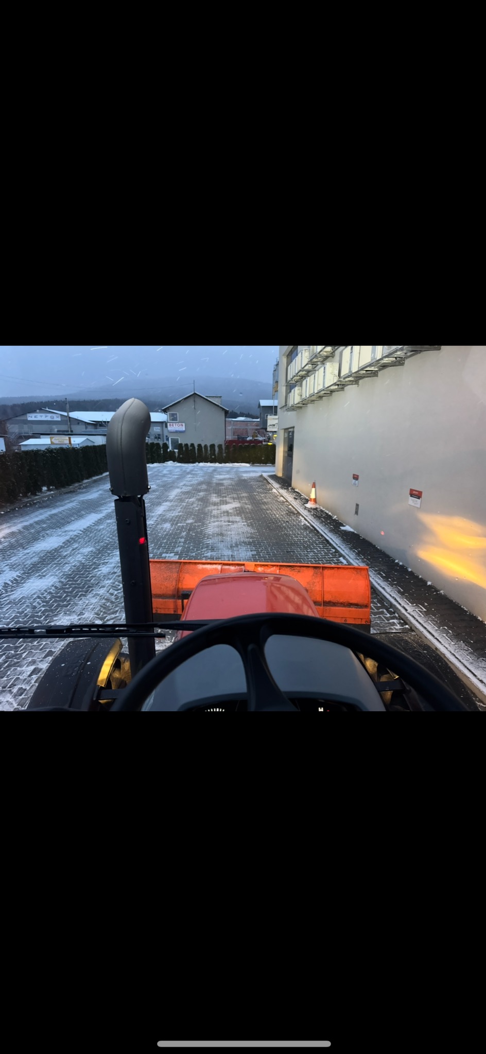 Widok z kabiny traktora odśnieżającego kostkę brukową w zimowy dzień. Budynki w tle, oświetlenie włączone. Perspektywa zza kierownicy.