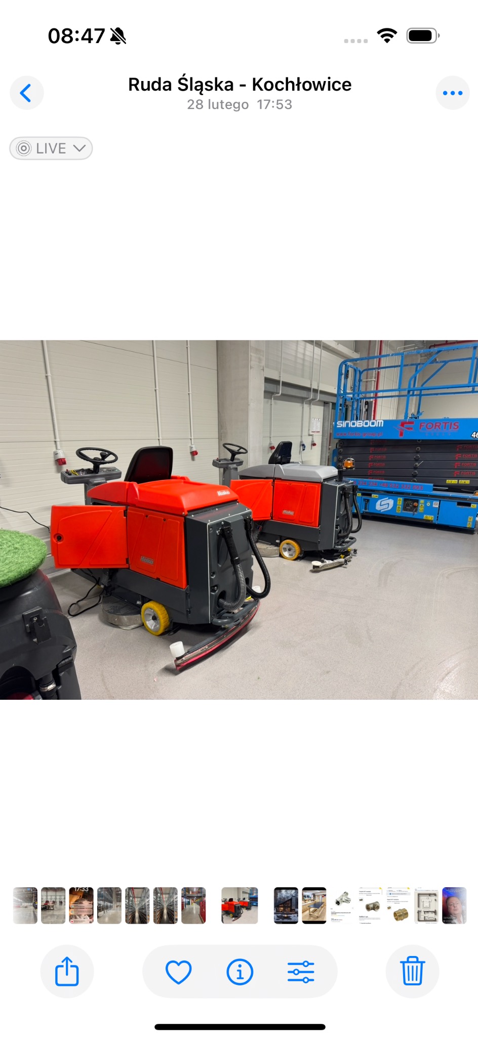 Dwie pomarańczowe maszyny czyszczące Hako w hali przemysłowej. W tle podnośnik Sinoboom Fortis. Widoczne ślady czyszczenia podłogi. Hala oświetlona lampami.