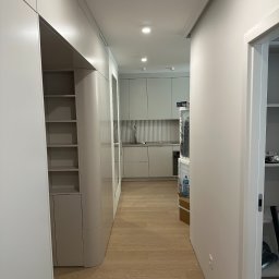 Mistre Group sp. z o.o. - Nowoczesne wykończenie wnętrza: korytarz z zabudową meblową w odcieniach szarości, drewniana podłoga i widok na kuchnię z ryflowanym panelem ściennym.