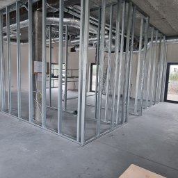 Mistre Group sp. z o.o. - Montaż szkieletu ścian działowych z profili stalowych w surowym wnętrzu. Na pierwszym planie narzędzia: wkrętarka, dalmierz i rękawice robocze.